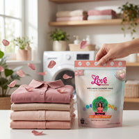 Stack of folded pink and beige clothes with a package of Love Brands Wellness Laundry Paks in a laundry room setting.